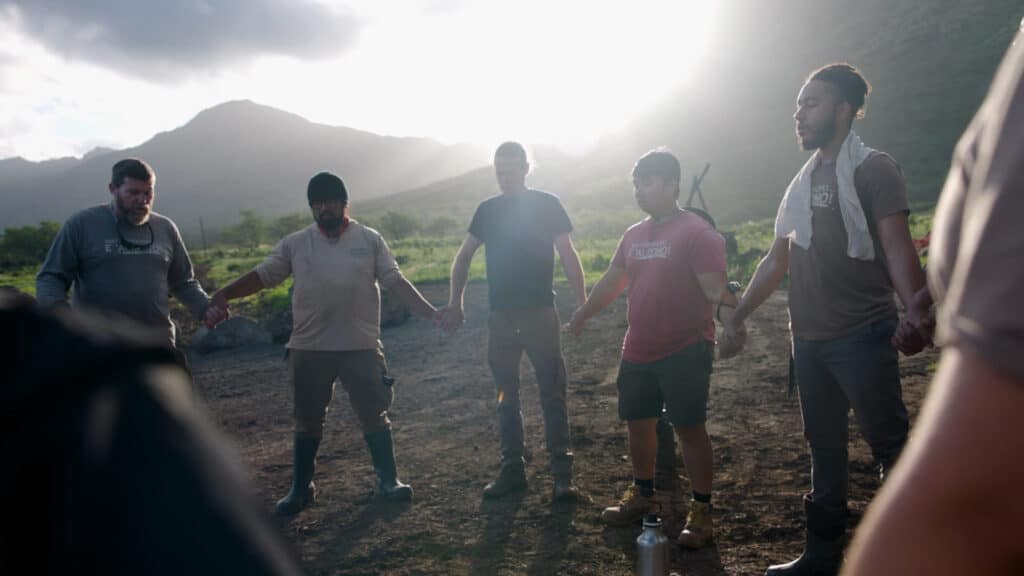 A group of people holding hands in a circle on a farm with mountains in the background.